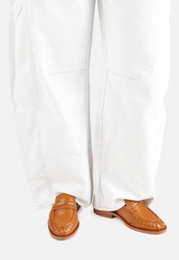 Mocasines de cuero marrón con un acabado texturizado, un acento metálico decorativo, combinados con pantalones de pierna ancha blancos con costuras visibles.
