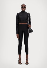 Black, long-sleeve cropped top featuring a high neck, paired with black, form-fitting leggings. Accessory includes a small black bag with chain details.