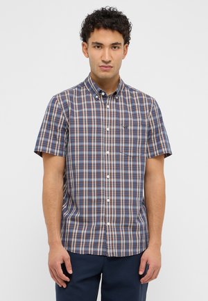 Young man wearing a short-sleeve button-up shirt with blue, white, and brown plaid pattern and navy pants on a plain background.