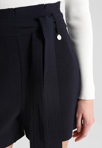 Shorts navy en tissu texturé, dotés d'une ceinture à nouer avec un accent métallique. Associés à un haut à manches longues côtelé blanc.