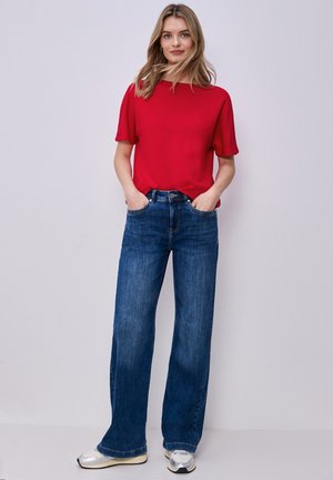 Jonge vrouw staat met handen in de zakken, draagt een rood topje met korte mouwen, blauwe wijd uitlopende jeans en zilveren sneakers tegen een effen achtergrond.