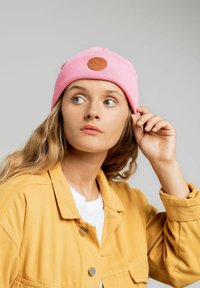 Różowa czapka typu beanie z prążkowaną teksturą i okrągłą brązową skórzaną łatą z przodu, noszona przez osobę w żółtej kurtce.