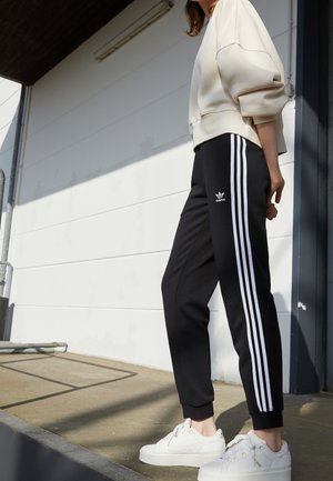 Osoba nosząca czarne spodnie dresowe Adidas z białymi paskami i białe trampki, stojąca na zewnątrz przy białej ścianie w świetle słonecznym.