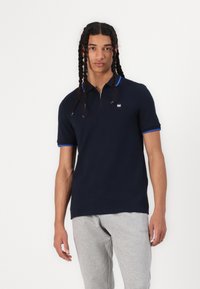 Polo shirt marine avec un col zippé, accents bleus sur le col et les bords des manches, en tissu de coton. Associé à un pantalon de survêtement gris.