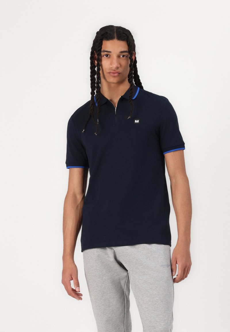 Polo shirt marine avec un col zippé, accents bleus sur le col et les bords des manches, en tissu de coton. Associé à un pantalon de survêtement gris.