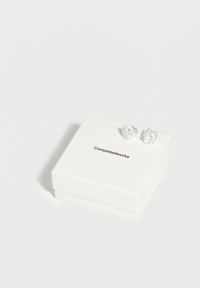 Ζευγάρι ασημένια μοτίβο σκουλαρίκια καρφωτά που ακουμπούν σε ένα λευκό κουτί κοσμημάτων με την επωνυμία Completedworks, σε απλό λευκό φόντο.