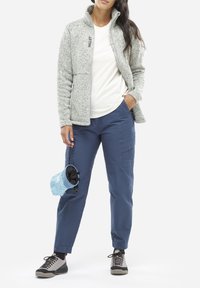 Veste en polaire gris clair avec col montant, associée à un t-shirt blanc et un pantalon cargo bleu. Chaussures noires et grises, ainsi qu'une pochette bleue à motifs.