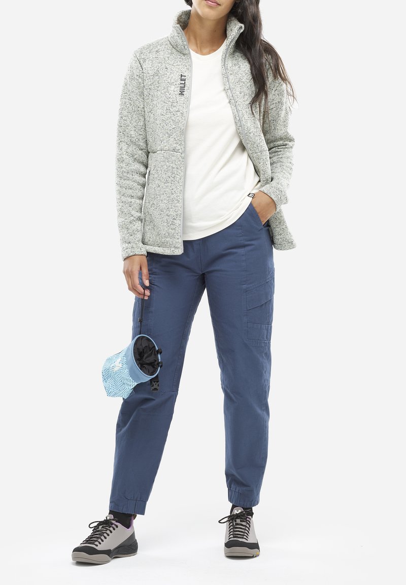 Veste en polaire gris clair avec col montant, associée à un t-shirt blanc et un pantalon cargo bleu. Chaussures noires et grises, ainsi qu'une pochette bleue à motifs.