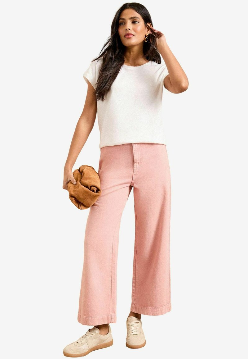 T-shirt blanc léger, ample, associé à un pantalon large taille haute rose pastel. Chaussures marron clair et un objet pliable beige en main.