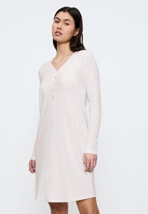 Kvinna klädd i en långärmad, ljusbeige, knälång klänning med V-ringning och fem knappar på bröstet, stående mot en enfärgad bakgrund.