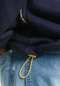 Navy Fleece Sweatshirt mit strukturierten Oberflächen, ausgestattet mit einem gelben Zugband, das am Saum verstellbar ist, getragen mit verwaschenen blauen Jeans.