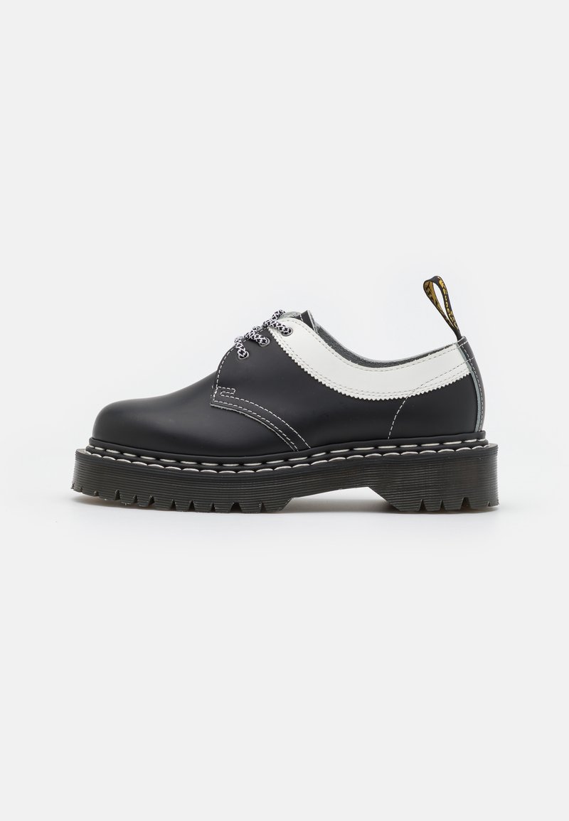Chaussure basse en cuir noir et blanc avec semelle épaisse crantée, lacets ronds noirs et blancs, et languette de traction à logo sur le talon.