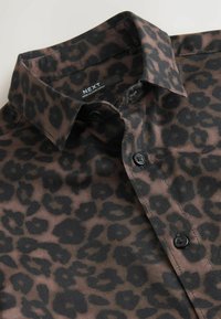 Chemise à boutons marron foncé avec un motif léopard noir et un col classique, étiquetée "NEXT Fabriqué au Bangladesh."