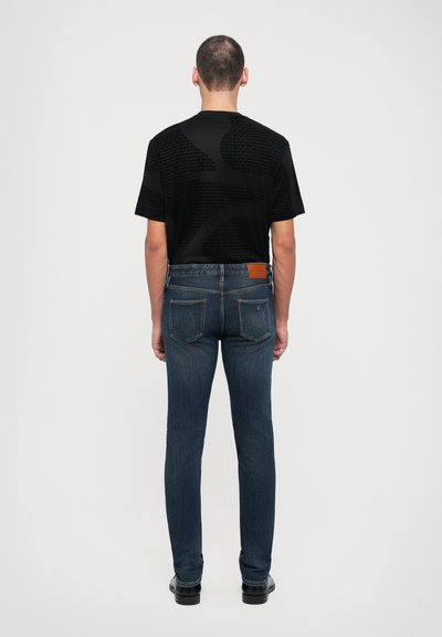 Camisa preta de manga curta com padrão texturizado nas costas, combinada com jeans de corte slim azul escuro com cinco bolsos e um rótulo de couro castanho.