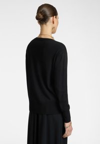 Donna con i capelli raccolti in uno chignon, che indossa un maglione nero a maniche lunghe e una gonna, accessoriata con gioielli d'oro, voltata di spalle contro uno sfondo bianco.