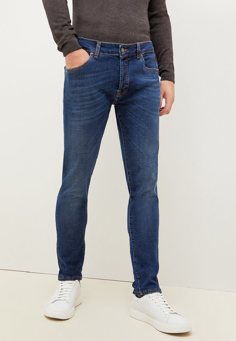 Jeans azul oscuro en un diseño ajustado, con un ligero desgaste, estilo tradicional de cinco bolsillos y acentos de costura en naranja.