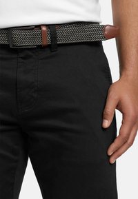 Cintura intrecciata con motivo a spina di pesce bianco e nero, con dettagli in pelle marrone e fibbia in metallo, indossata con pantaloni neri.