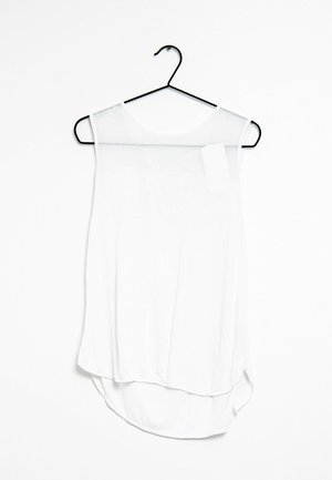 Débardeur blanc sans manches en tissu léger, avec un ourlet incurvé, un col arrondi et une section supérieure transparente. Accroché à un crochet noir.