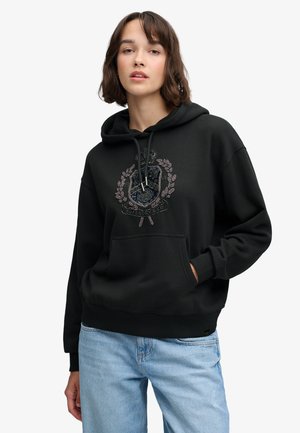 Jeune femme portant un sweat à capuche noir avec un blason brodé, associée à un jean bleu clair, debout devant un fond blanc.