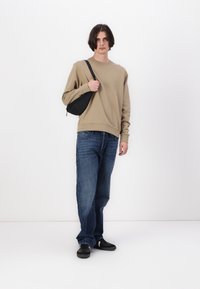 Beige színű pulóver hosszú ujjakkal, kék, laza szabású farmer és fekete sportcipő. A modell egy kis fekete válltáskát tart, ami pánttal rendelkezik.