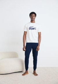 Tricou din bumbac alb cu un grafic de crocodil bleumarin, asociat cu pantaloni tapering bleumarin. Fundal deschis, neutru.