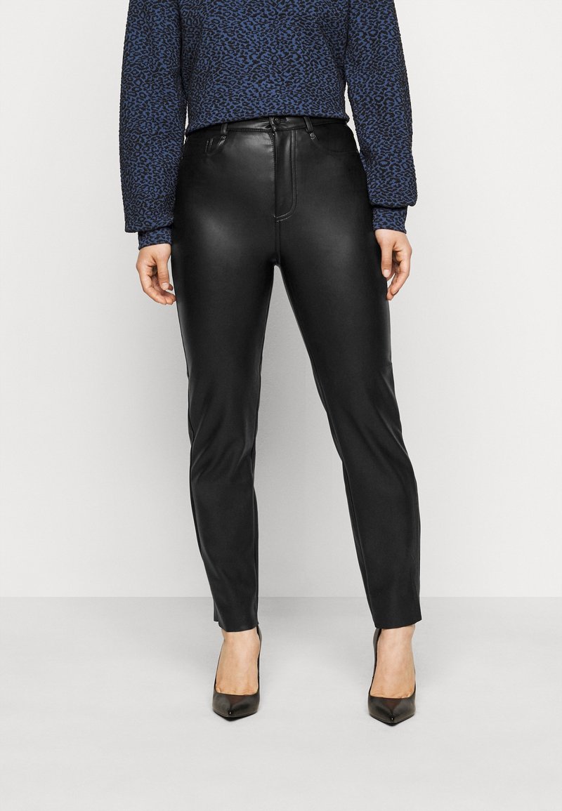 Personne portant un pantalon en cuir noir à coupe slim, un haut à manches longues sombre à motifs, et des escarpins noirs pointus, se tenant devant un fond blanc.