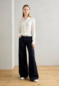 Chemise blanche à manches longues avec texture côtelée, associée à un jean évasé foncé. Des baskets blanches complètent la tenue. Bijoux en argent simples présentés.