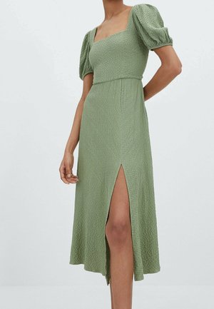 Vrouw die een midi-jurk draagt in een gestructureerde saliegroene kleur met pofmouwen en een hoge split aan de voorkant, staand tegen een effen lichte achtergrond.