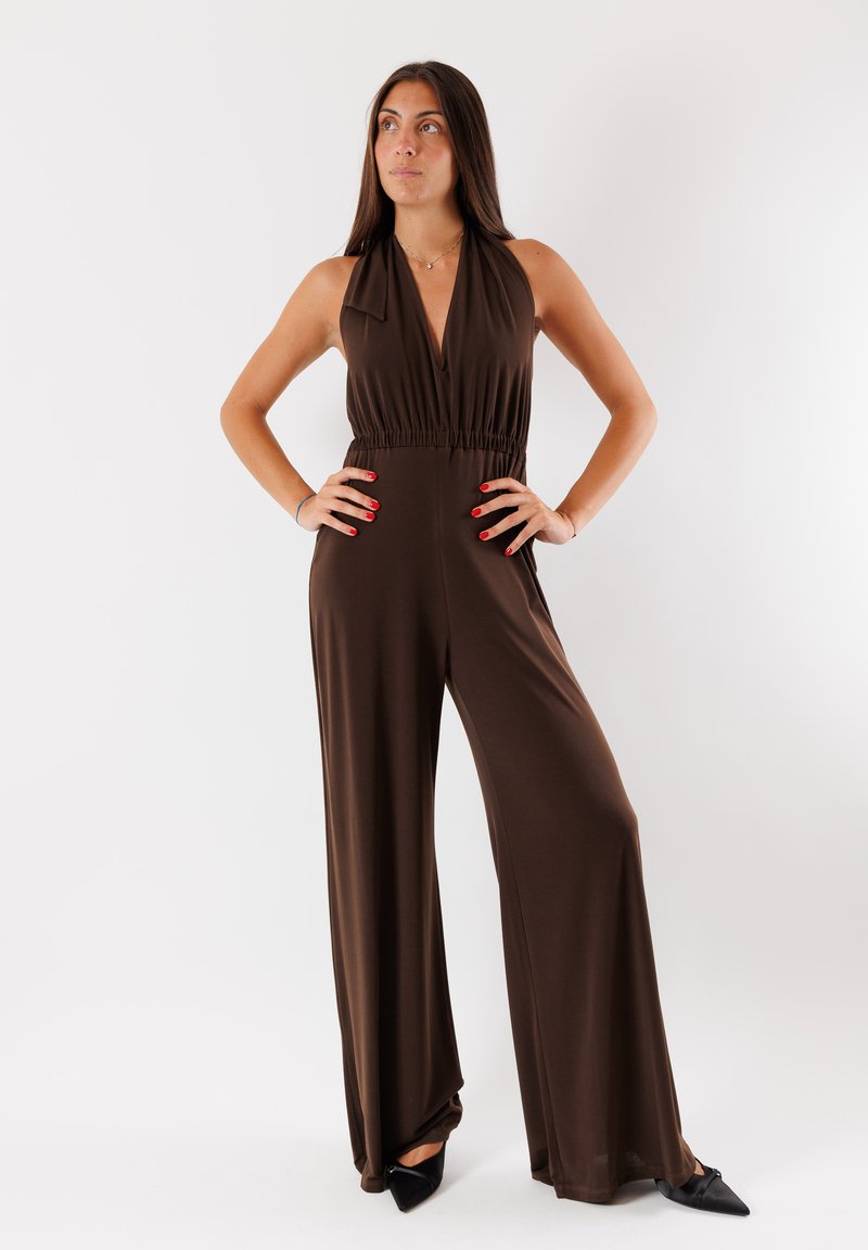 Jumpsuit marrone con scollo a V profondo, vita elastica e gambe larghe; abbinato a tacchi neri a punta su uno sfondo bianco.