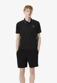 Svart polo skjorta med subtil geometrisk mönster, gula accenter på kragen och en broderad logotyp. Matchad med svarta shorts.