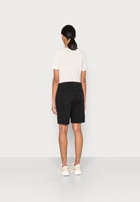 Žena stojící zády, oblečená v lehce pruhované krátkolemé košili, černých kraťasech do kolen a béžových teniskách na bílém pozadí.