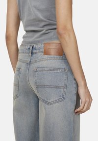 Niebieskie dżinsy o luźnym kroju, z brązową skórzaną metką na pasku i kontrastowym przeszyciem. Widać tylne kieszenie.