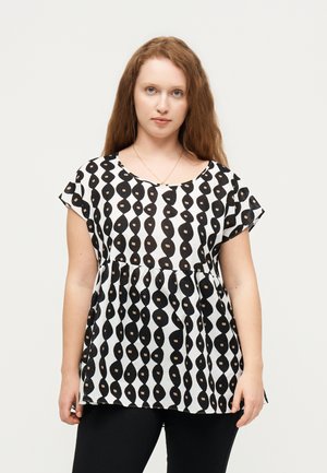 Hosszú vörös hajú nő, fekete-fehér mintás rövid ujjú blúzban és fekete nadrágban, előre néz egy egyszínű háttér előtt.