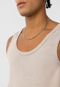 Beige geribbelde tanktop met ruwe randen, met een ronde halslijn; een zilveren ketting ligt op het sleutelbeen; gladde textuur.