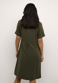 Vestido de lino verde oliva con mangas cortas, un cuerpo ajustado y un diseño de espalda plisada, que cae justo por encima de la rodilla. Textura suave, silueta simple.