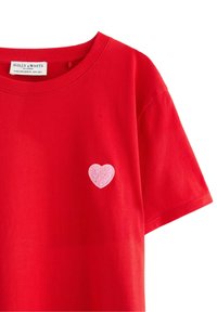 Czerwona bawełniana koszulka z luźnym krojem, z różowym haftowanym sercem na lewej stronie klatki piersiowej. Okrągły dekolt i krótkie rękawy.