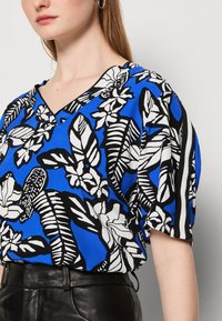 Top azul con un atrevido patrón floral en blanco y negro, con escote en v y mangas cortas, combinado con pantalones de cuero negros.