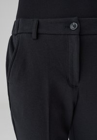 Pantaloni neri con tessuto a trama, dotati di chiusura con un bottone, passanti per cintura e tasche laterali. Il tessuto è liscio e leggero.