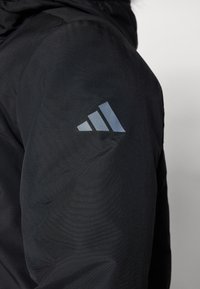 Μαύρο μπουφάν με λεία, υφασμάτινη υφή και ανακλαστικό γκρίζο τριγωνικό λογότυπο στο αριστερό μανίκι.