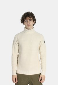 Maglione a collo alto color crema realizzato in tessuto a coste, con maniche lunghe e una silhouette aderente, impreziosito da una toppa con logo sul braccio.