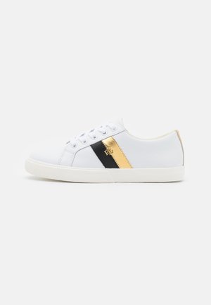 Zapatilla blanca de perfil bajo con cordones blancos, franjas laterales metálicas negras y doradas, y suela plana de goma.