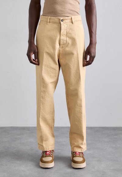 Pantalons en coton beige clair avec une coupe décontractée, dotés de poches frontales, d'une fermeture à bouton, portés avec des baskets marron et roses.