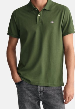 Mężczyzna w oliwkowej, krótkiej koszulce polo z małym logo na piersi, w połączeniu z wyblakłymi czarnymi dżinsami, z ręką w kieszeni.