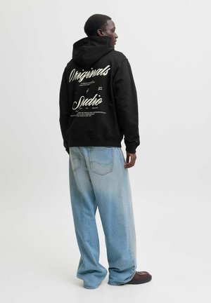 Homme portant un sweat à capuche noir avec texte blanc et jean large bleu clair, debout et regardant sur le côté sur un fond uni.