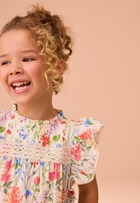 Florale jurk van lichtgewicht stof met ruffle mouwen, voorzien van een witte kantafwerking op de borst. Kleuren zijn roze, blauw, groen en geel.