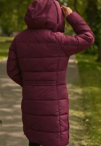 Manteau en duvet long bordeaux avec une capuche, comportant des panneaux matelassés horizontaux et des poches latérales. Texture lisse avec une silhouette ajustée.