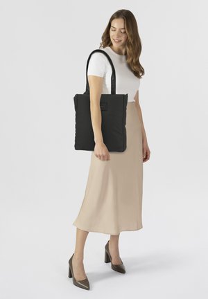 Schwarze Nylon-Tasche mit zwei langen Griffen, die eine strukturierte Oberfläche und ein rechteckiges Schild auf der Vorderseite aufweist, gehalten von einer Person in einem weißen Oberteil und einem beigen Rock.