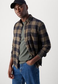 Cămașă cu carouri din flanel în nuanțe de marron și bleumarin, asortată cu un tricou gri cu guler rotund și blugi. Prezintă un sistem de închidere cu butoni și mâneci rulate.