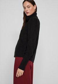 Melnas ribotas adītas turtlenecks džemperis ar garām piedurknēm, veidots ar brīvu piegriezumu, apvienots ar bordo biksēm.