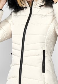 Giacca imbottita color crema con forma aderente, zip frontale e dettagli in zip neri a contrasto. Presenta un cappuccio foderato in pelliccia e pannelli laterali con texture.